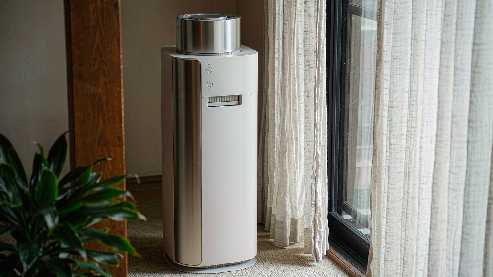 A tall cylindrical air purifier beside a bedroom window with sheer curtains and a potted plant in soft morning light