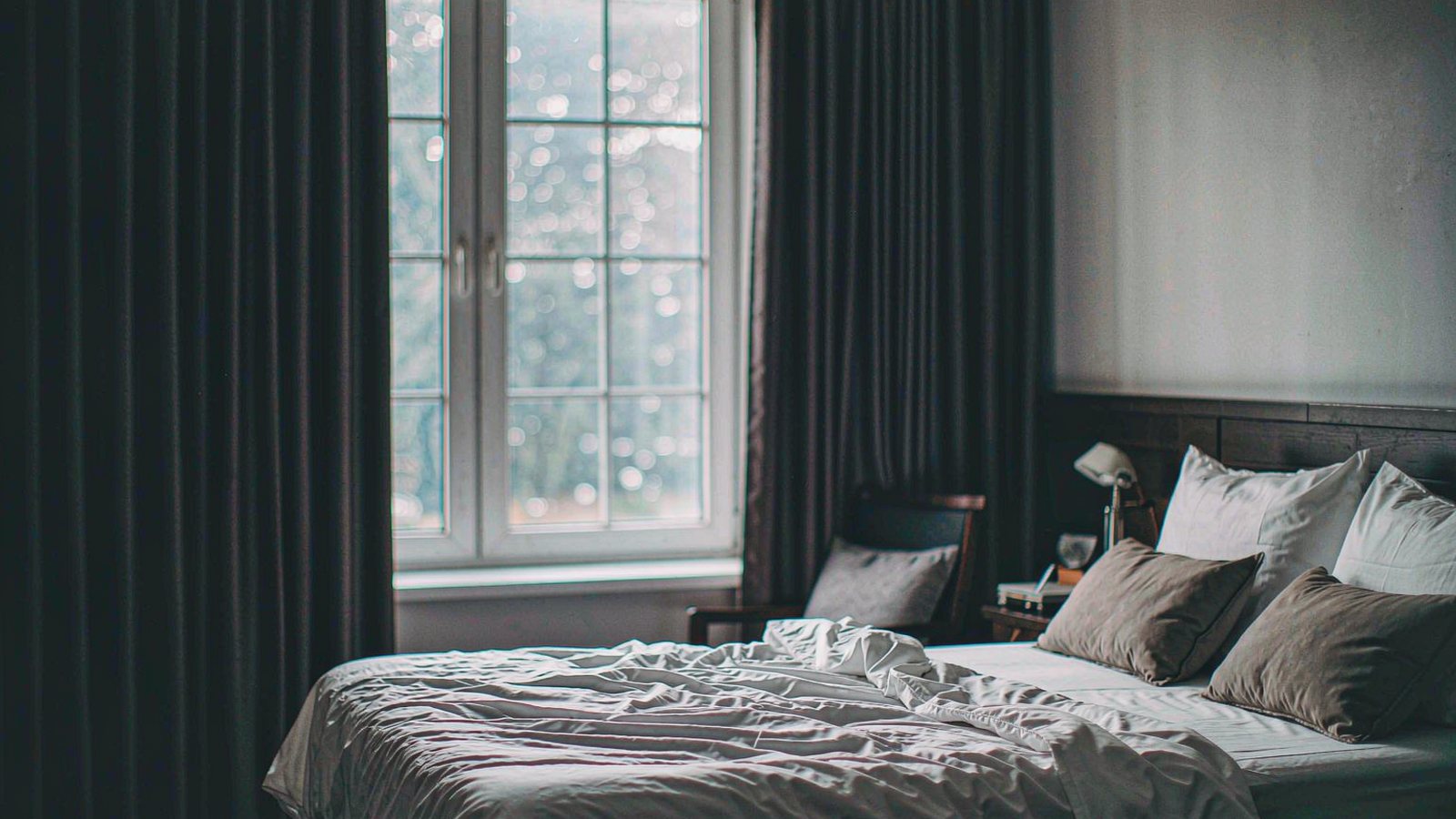 Heavy dark curtains drawn across a bedroom window with a thin sliver of daylight at the edge and a calm dim interior