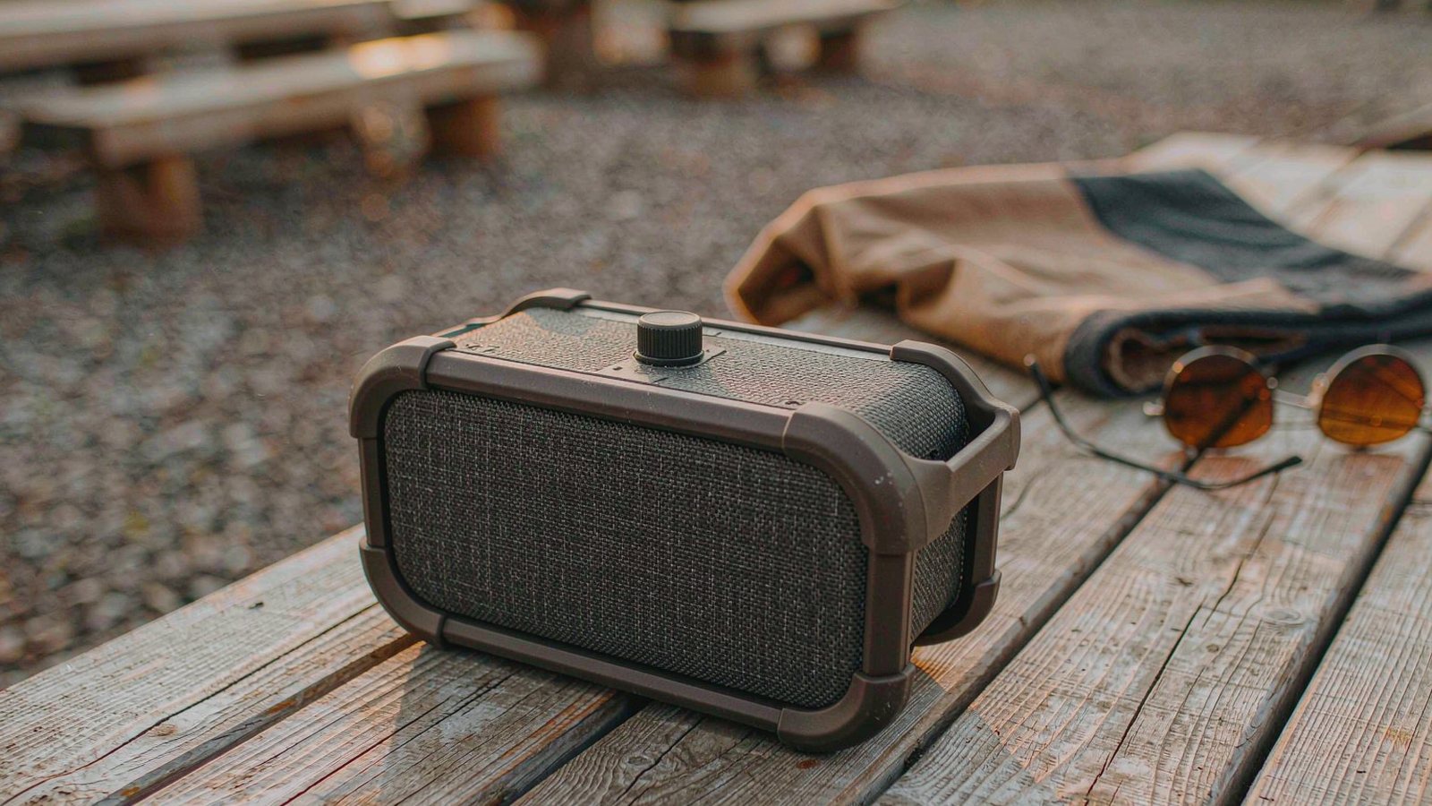 A rugged portable bluetooth speaker sitting on a weathered wooden outdoor table with a picnic blanket and sunglasses in golden hour light