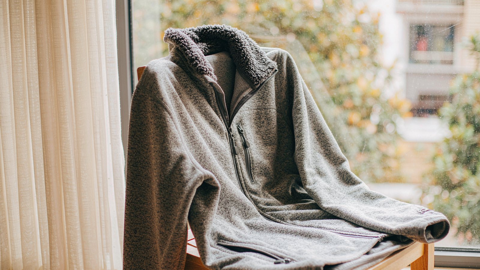 A fleece quarter-zip pullover draped over a chair by a window with soft autumn light and leaves outside