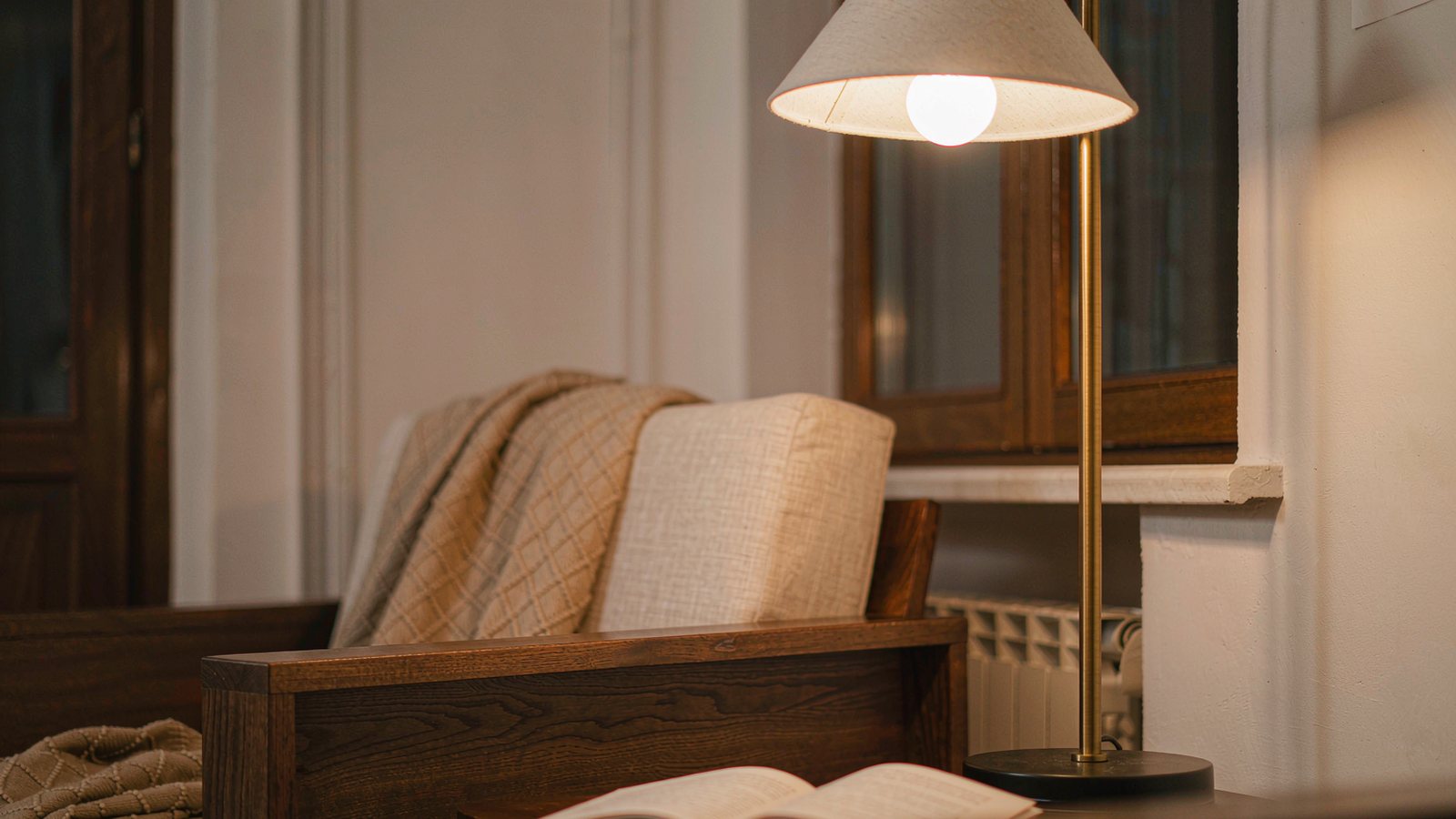 A warm brass floor lamp beside a linen armchair with an open book and a folded blanket in cosy evening light