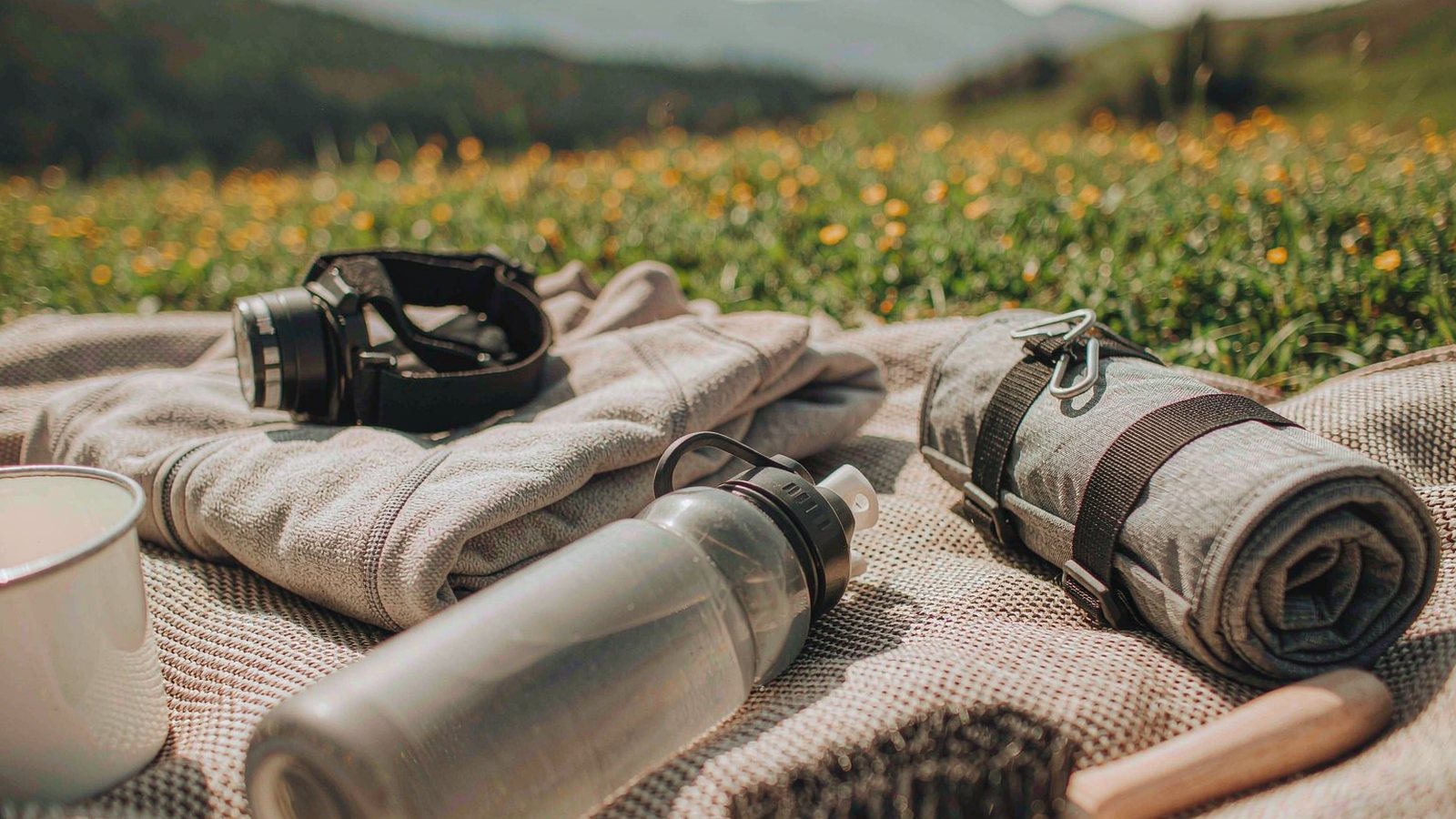Outdoor gear including socks, a headlamp and a water bottle laid out on a picnic blanket in a meadow with mountains behind