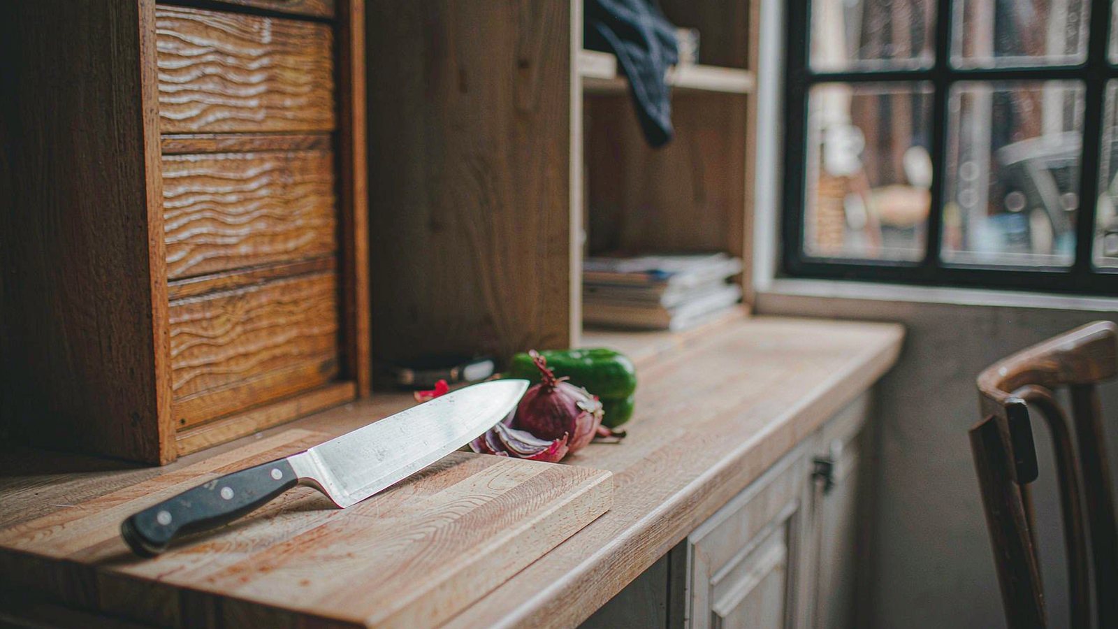 A chef's knife resting beside a wooden cutting board with fresh vegetables on a warm kitchen counter in natural light