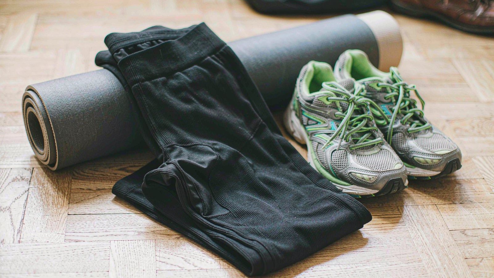 Black leggings folded on a light oak floor beside running shoes and a rolled yoga mat in soft morning light