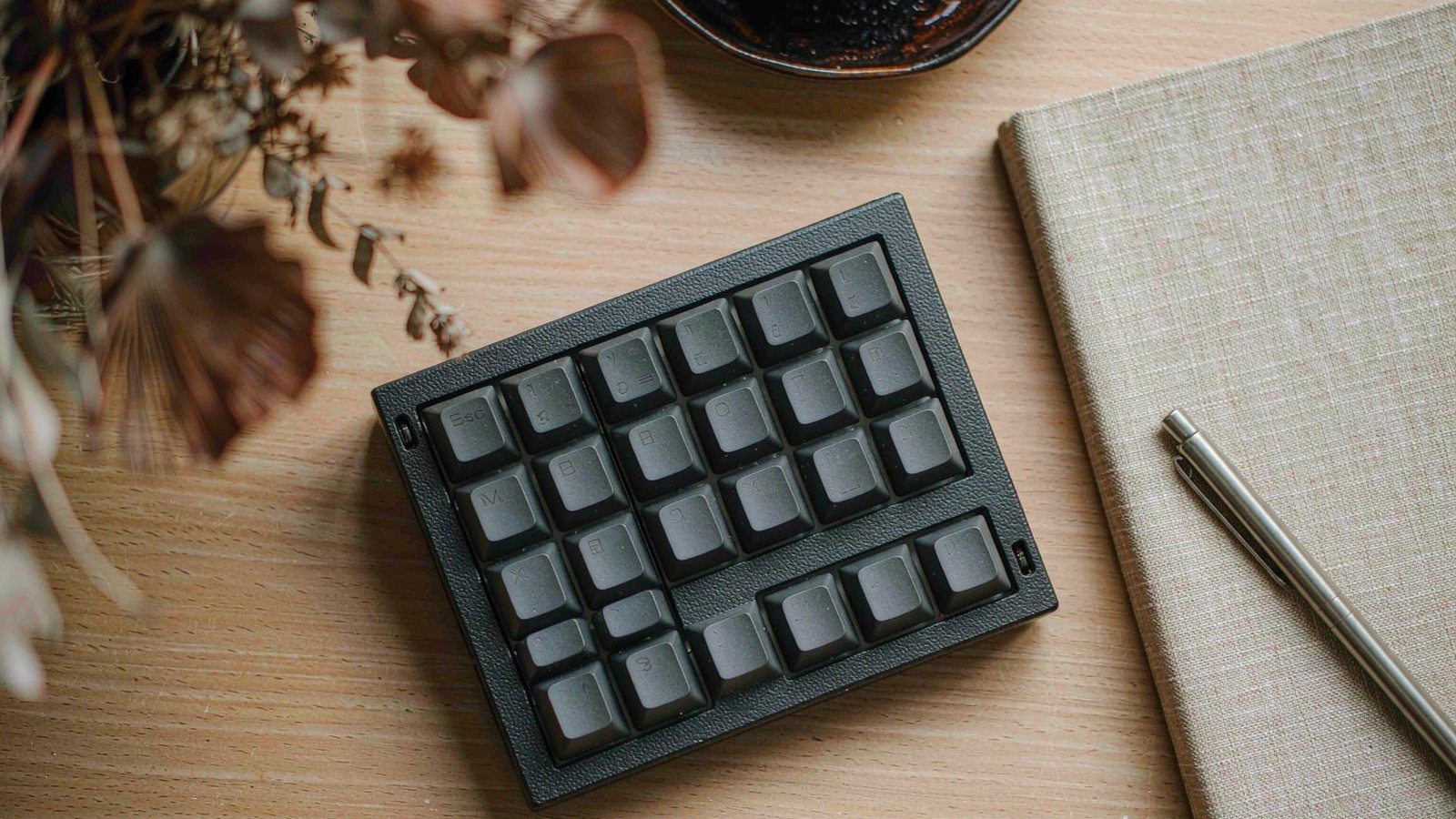 An overhead view of a compact mechanical keyboard with plain keycaps on a walnut desk beside a notebook and a pen in soft daylight