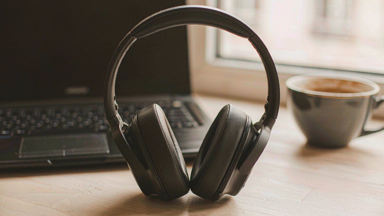 Over-ear headphones resting on a desk beside a laptop and a cup of coffee in a sunlit home office
