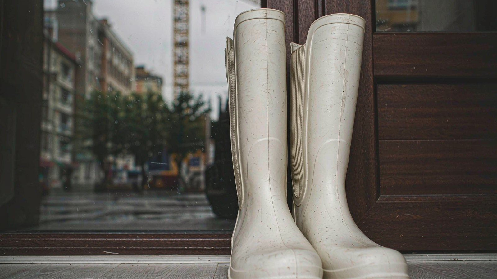 Tall rubber rain boots standing by a doorway with a wet street reflected in the glass behind, photographed in overcast light.