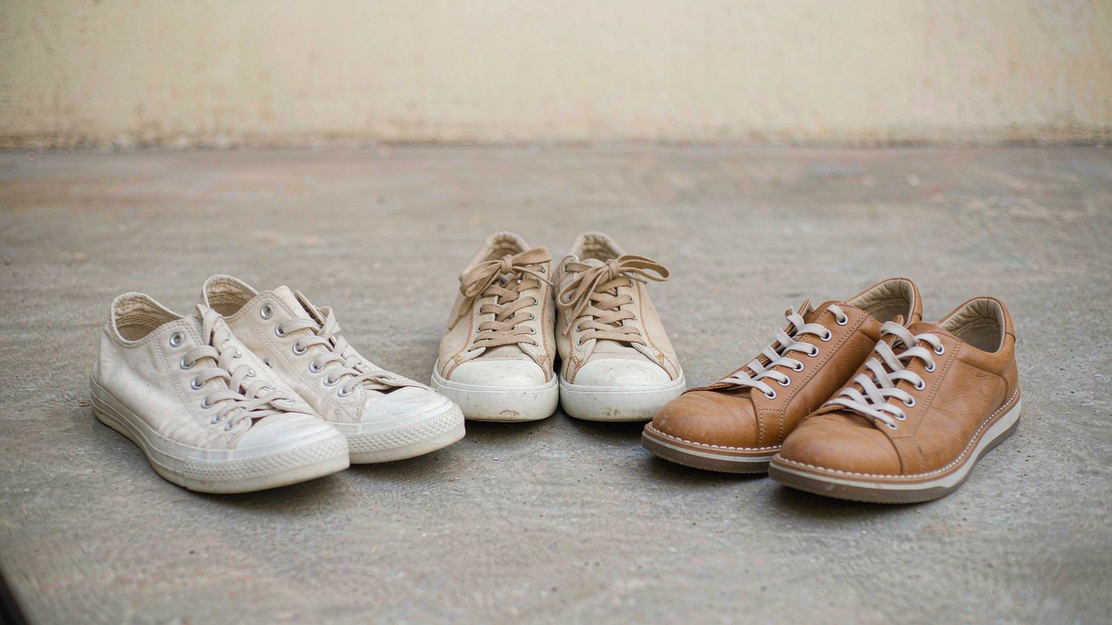 Three pairs of neutral sneakers lined up on a concrete floor against a plain pale wall in soft daylight