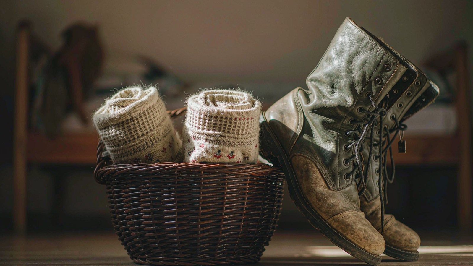 Rolled wool socks in a woven basket on a wooden floor beside a pair of worn leather boots in warm light