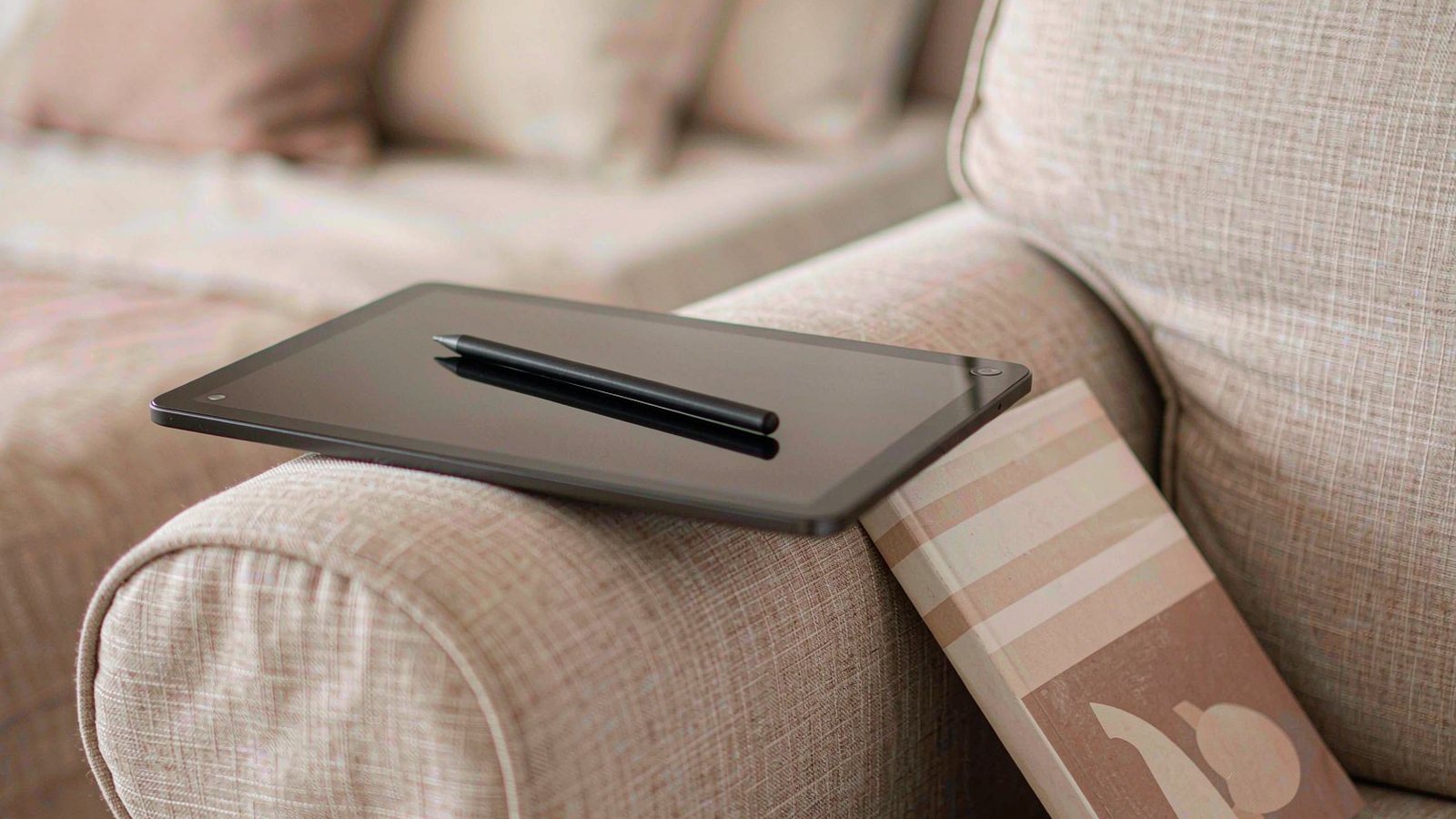 A tablet resting on the arm of a linen sofa with a stylus and a paperback book beside it in soft natural light