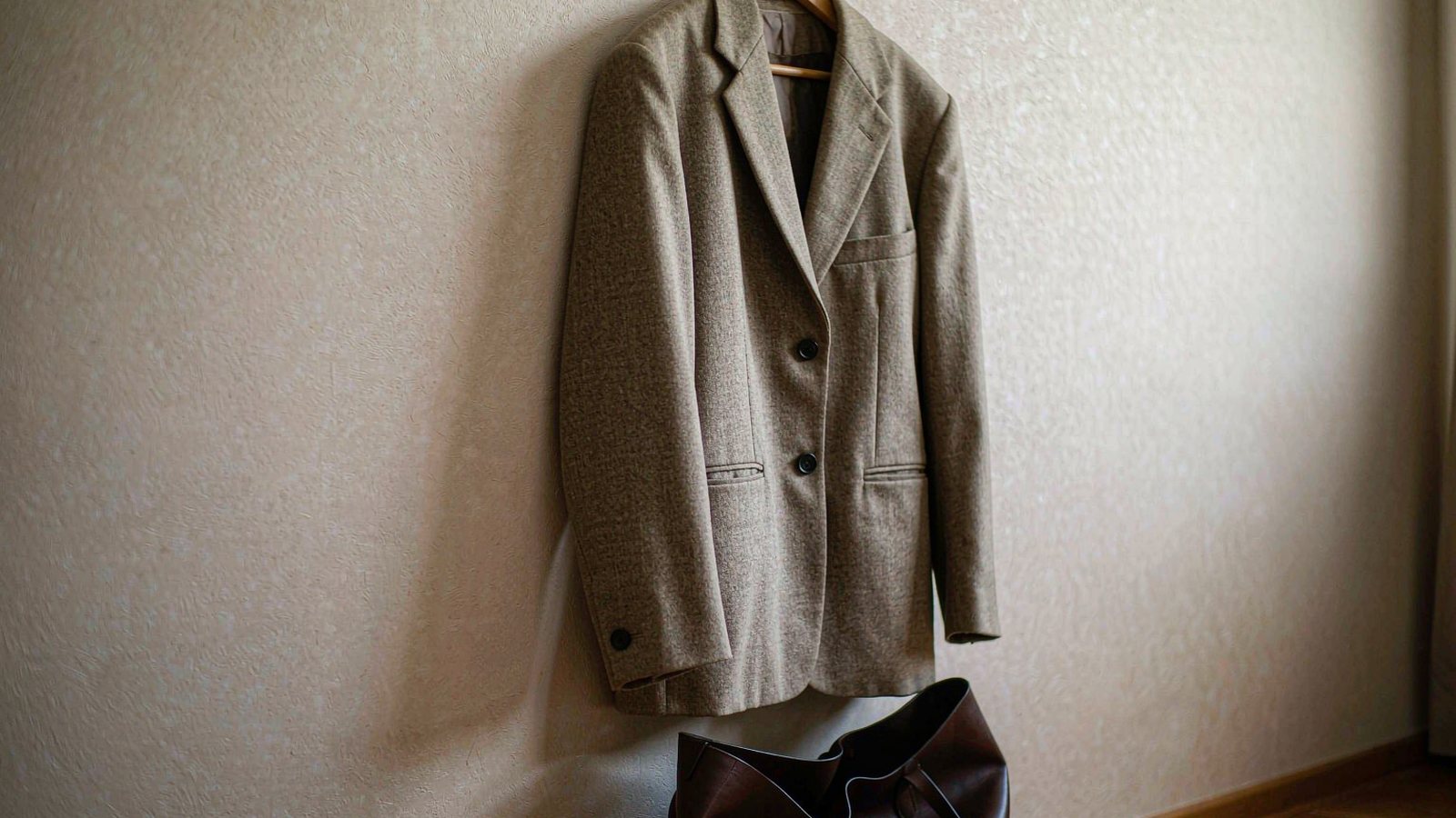 A structured neutral blazer on a wooden hanger against a warm plaster wall with a simple canvas tote bag nearby in soft daylight